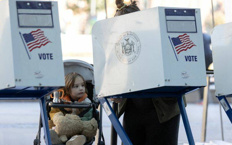 Una niña en un cochecito observa a una mujer emitir su voto. La mujer está protegida por una pantalla de privacidad que dice vote con la bandera estadounidense representada en ella.